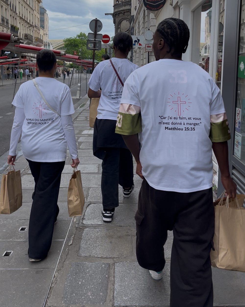 Trois bénévoles marchent dans Paris, sacs solidaires à la main, de dos avec le verset Matthieu 25:35 sur leur t-shirt